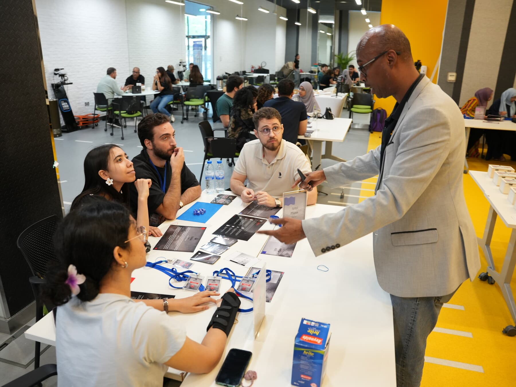 A facilitator leaning over one team's table mid-round to answer a question, the rest of the room playing on behind him.