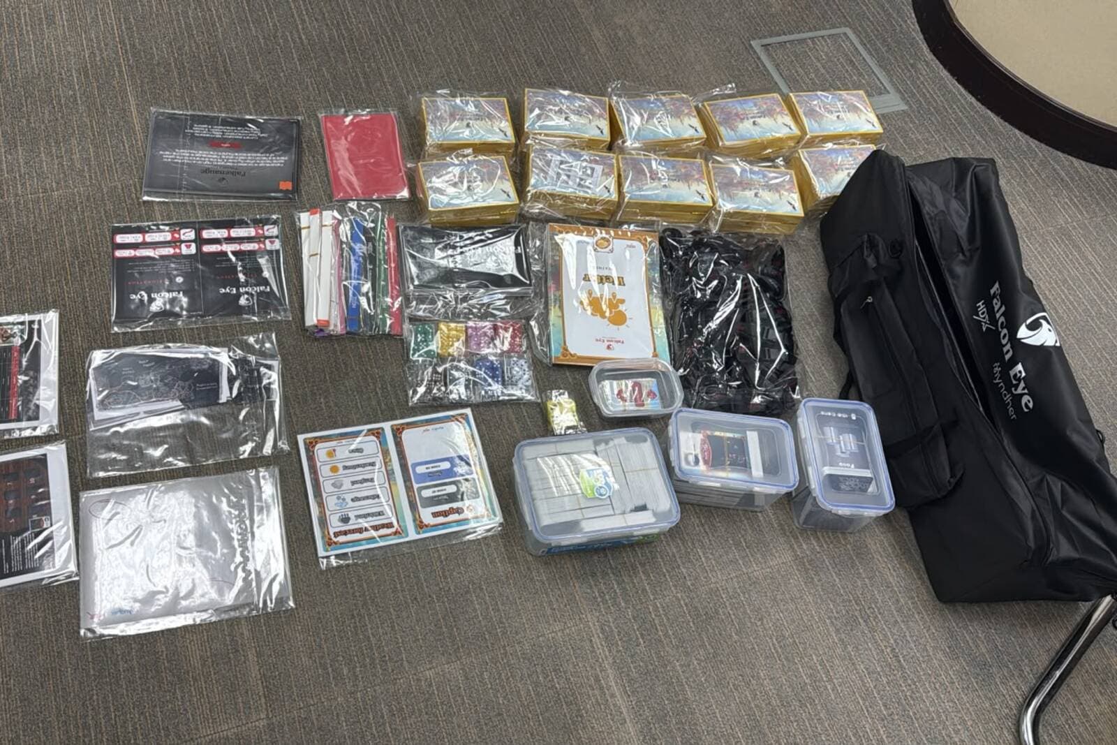 A complete Falcon Eye kit unpacked across a table: bagged card decks, resource tokens, printed reference cards, four lidded component cases and the branded carry bag it all travels in.