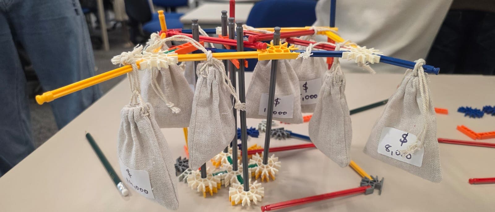 A team's Rooted structure part-way through a round: weighted cloth revenue pouches, each hand-marked with a dollar value, hanging from K'NEX rods.
